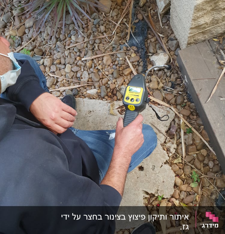 אדם מחזיק מכשיר איתור צינורות על קרקע עם אבנים